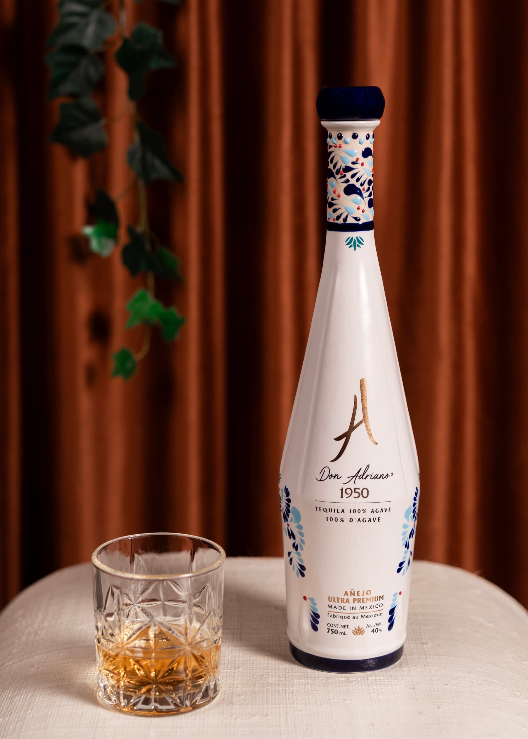 Don Adriano 1950 Añejo Ultra Premium in its blue-and-white ceramic bottle, beside a cut-crystal glass of tequila, set against a rust-coloured curtain